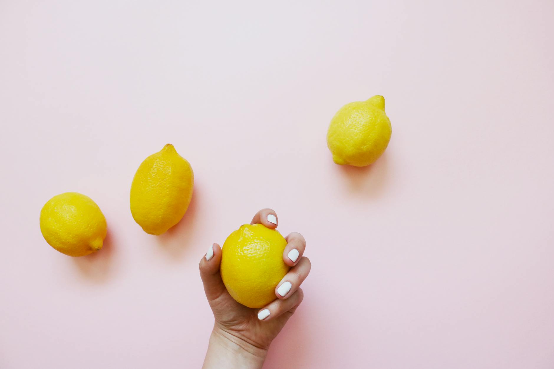 Hand holding a bright yellow lemon on a soft pink background with additional lemons nearby