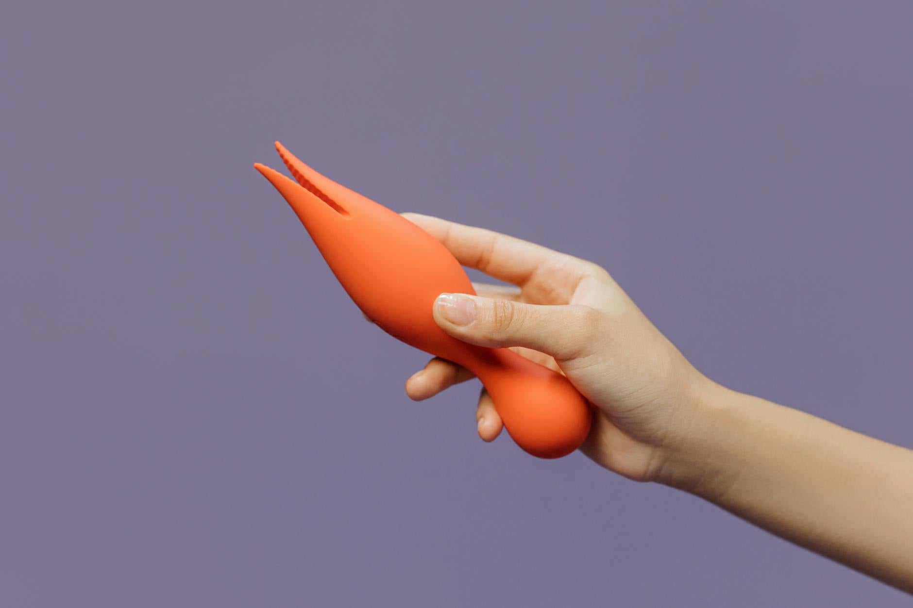 A close-up of a hand holding an orange vibrator against a minimalistic purple backdrop showcasing modern sensuality
